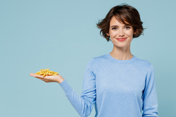 Young smiling woman wearing casual sweater looking camera hold in palm hand fastfood french fries potato isolated on plain pastel light blue background studio portrait. People lifestyle food concept.
