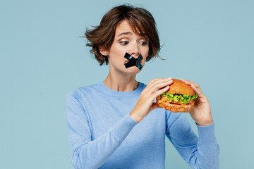 Young wistful woman with mouth sealed with tape in sweater look camera hold burger want to eat isolated on plain pastel light blue background studio portrait. People lifestyle diet junk food concept.