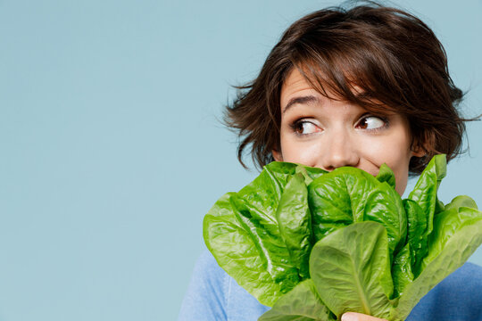 Close Up Young Woman 20s In Sweater Hold Cover Mouth With Green Lettuce Leaves Look Aside On Workspace Isolated On Plain Pastel Light Blue Background Studio Portrait. People Lifestyle Food Concept