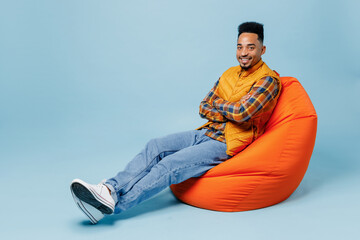 Full size body length smiling young black man 20s years old wears yellow waistcoat shirt sit in bag chair hold hands crossed look camera isolated on plain pastel light blue background studio portrait.