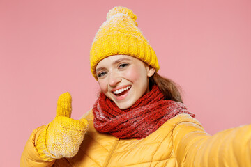 Close up fun young woman 20s years old wears yellow jacket hat mittens do selfie shot pov on mobile phone showing thumb up like gesture isolated on plain pastel light pink background studio portrait.