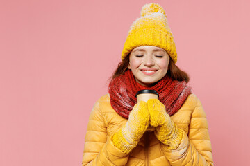 Young woman 20s wears yellow jacket hat mittens keep eyes closed hold takeaway delivery craft paper brown cup coffee to go smell sniff isolated on plain pastel light pink background studio portrait.