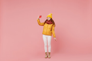 Full size body length young woman 20s wear yellow jacket hat mittens doing selfie shot on mobile cell phone post photo on social network isolated on plain pastel light pink background studio portrait