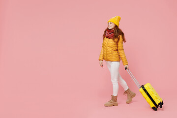 Full size body length happy young woman 20s years old wears yellow jacket hat mittens look aside hold push suitcase bag walking stepping isolated on plain pastel light pink background studio portrait.