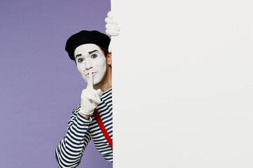 Young mime man with white face mask wears striped shirt beret look out from behind white wall say hush be quiet with finger on lips shhh isolated plain pastel light violet background studio portrait.