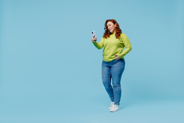 Full body smiling fun young chubby overweight plus size big fat fit woman wearing green sweater hold use mobile cell phone isolated on plain blue background studio portrait. People lifestyle concept.