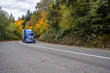 Fototapeta na wymiar Bright blue fresh big rig semi truck transporting frozen cargo in reefer semi trailer driving on the curved road through the autumn forest