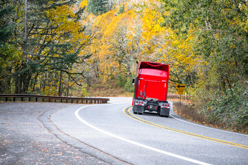 Bright red big rig semi truck with high cab driving without semi trailer on the winding mountain road with autumn yellow trees forest