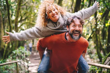 Fotobehang Overjoyed adult couple have fun together at outdoor park in leisure activity. Man carrying woman in piggyback and laugh a lot. Love and life mature people lifestyle concept. Enjoying vacation nature © simona