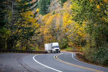 Powerful white big rig semi truck transporting goods in dry van semi trailer climbing up hill on the winding mountain road with autumn forest on the rocks