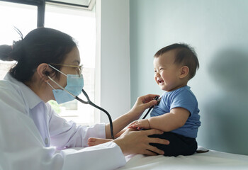 Asian female doctor wearing face mask examining little cute Baby boy with stethoscope