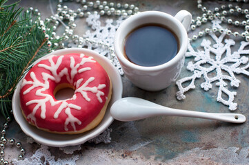Donut with pink icing and white icing snowflake with a cup of coffee and knitted snowflakes, white spoon, silver beads, fir branch on a grey background
