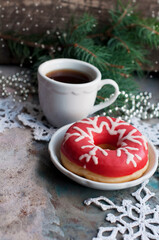 Donut with pink icing and white icing snowflake with a cup of coffee and knitted snowflakes, white spoon, silver beads, fir branch on a grey background
