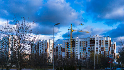 An airplane is flying in the cloudy autumn sky over new buildings and a construction crane