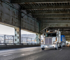 White classic big rig bonnet semi truck with additional lights transporting covered cargo on flat bed semi trailer running on the two level arch transportation Fremont bridge