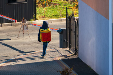 A courier with a large yellow bag on his back walks to a closed metal gate into the protected area. Delivery of goods and safety during the coronavirus pandemic