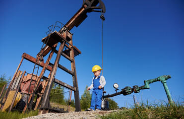 Bottom view of small young power engineer near oil rig. Boy in overalls against the blue sky, petroleum pipeline and pump jack for extracting natural fuel.