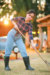Young asian woman gardener in red plaid shirt smiling woman wearing boots black and holding rake with pile of autumn eaves and hay stack.