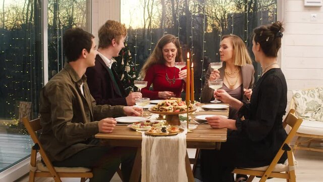 Friends celebrating new year, sitting at dining table. Young people drinking champagne. Diverse happy students men and women during christmas party, festive dinner at home.