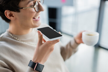 Smartphone with blank screen in hand of smiling businessman with cup recording voice message in office.