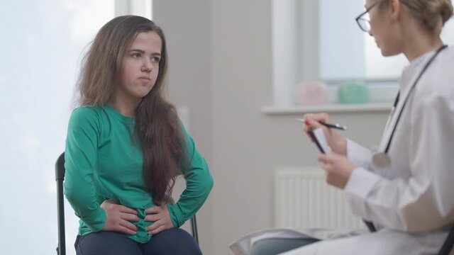 Female patient talking complaining abdominal pain symptoms of indigestion. Caucasian little person consulting professional doctor in eyeglasses in hospital. Illness and medicine concept