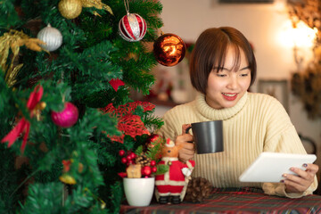 Beautiful woman in sweater sitting on chair to looking on tablet and drinking coffee cup near christmas tree