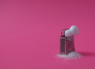 Half a white heart with grated pieces and a small grater on a pink background. Text space. Broken...