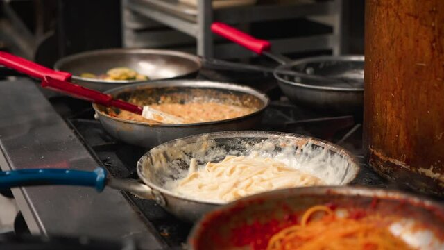Chef tosses to incorporate fresh pasta in alfredo cream sauce over Italian restaurant kitchen stove, pans with diffrent sauces lined up on prep stove, slow motion 4K