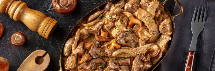Mushroom beef stroganoff panorama. Meat with champignons, boletus and ceps in a creamy sauce, in a skillet, shot from the top on a black background
