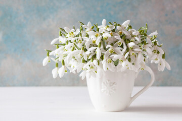 A bouquet of white spring flowers of snowdrops in a mug on a table against the background of a decorative wall, blur, selective focus.