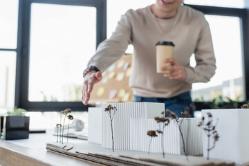 Cropped view of blurred businessman holding paper cup and pointing at model of building in office.