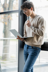 Young businessman holding pen and laptop near window in office.