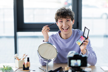 Happy transgender person applying eye shadow and looking a camera near decorative cosmetics and digital camera.