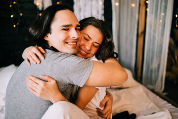 Fototapeta premium Lovely couple hugging on their bed decorated with garland lights at home indoors in the bedroom