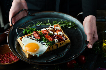 Sandwich with egg, cheese, salted fish and arugula on a plate in the hands of a chef on a rustic wooden background. Belgian waffles. On a black background.