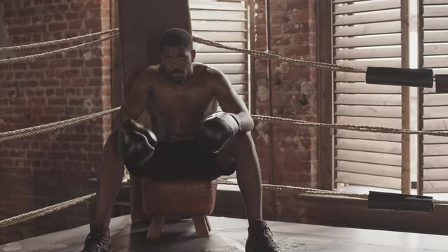 Slowmo portrait of shirtless Black man in boxing gloves looking at camera sitting in corner at boxing ring resting before fight
