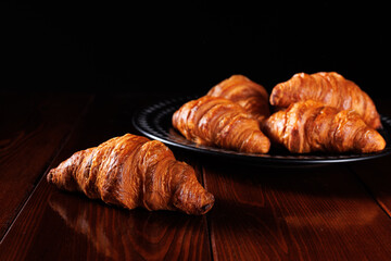 Beautiful baked croissants are served on a black dish on a wooden table. Angle view. Selective focusing.