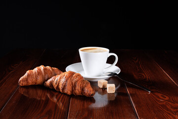 Two fresh croissants with a cup of coffee lie on a wooden table on a black background. Place for your text.