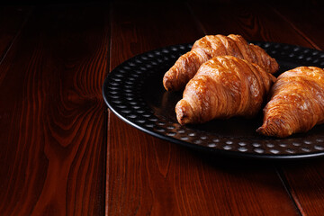 Fresh croissants lie on a black ceramic plate on a wooden table. Place for your text.