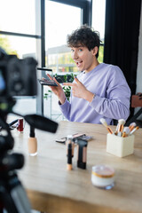 Excited transgender makeup artist pointing at eye shadows near cosmetics and digital camera.