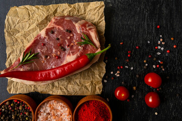 Raw piece of ribeye beef meat on craft paper on a wooden table stacked tomatoes garnished with hot peppers ,spice and himalayan salt. Fresh food. Everything for a barbecue.
