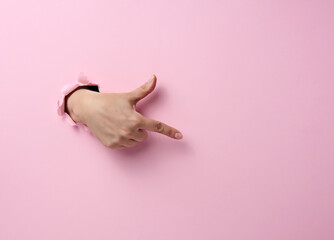 female hand sticks out of a torn hole in a pink paper background, part of the body points with the...