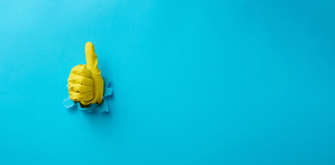 a hand in a yellow household rubber glove sticks out of a torn hole and shows a gesture okay, thumb up. Blue background