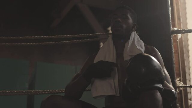 Medium slowmo portrait of tired African-American male boxer wiping sweat with towel sitting in rings corner resting after fight