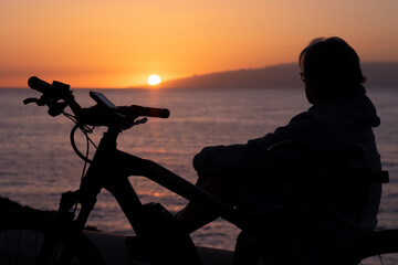 Naklejka premium Silhouette of woman with electric bicycle sitting after cycling along the sea looking at sunset. Orange-black background