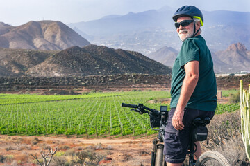 Old senior man enjoying outdoor excursion with bicycle. Elderly bearded man wearing sport helmet in countryside with vineyard and mountain