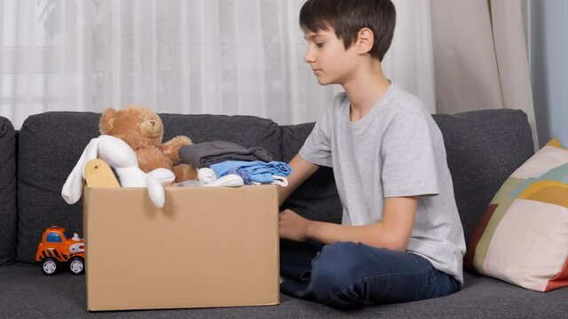 Boy preparing clothes and toys for charity donation. Kid sorting and packing kid toys, children clothes into cardboard box at home