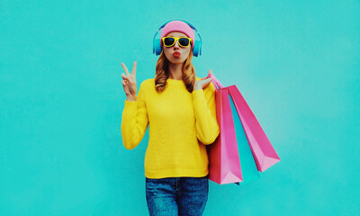 Fashionable portrait of stylish young woman listening to music in headphones with shopping bags posing wearing a yellow knitted sweater, pink hat on blue background