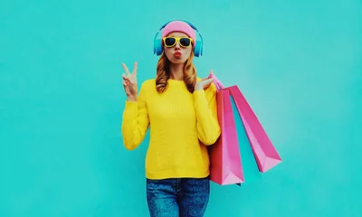 Photo sur Aluminium brossé Magasin de musique Fashionable portrait of stylish young woman listening to music in headphones with shopping bags posing wearing a yellow knitted sweater, pink hat on blue background