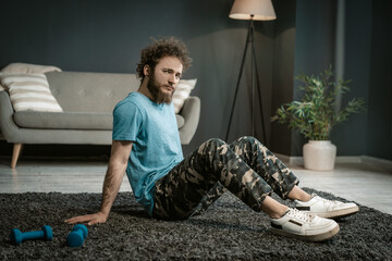 Man in a Blue T-shirt Sits on a Carpet Next to his Blue Dumbbells and Gazes at the Camera. Medium...